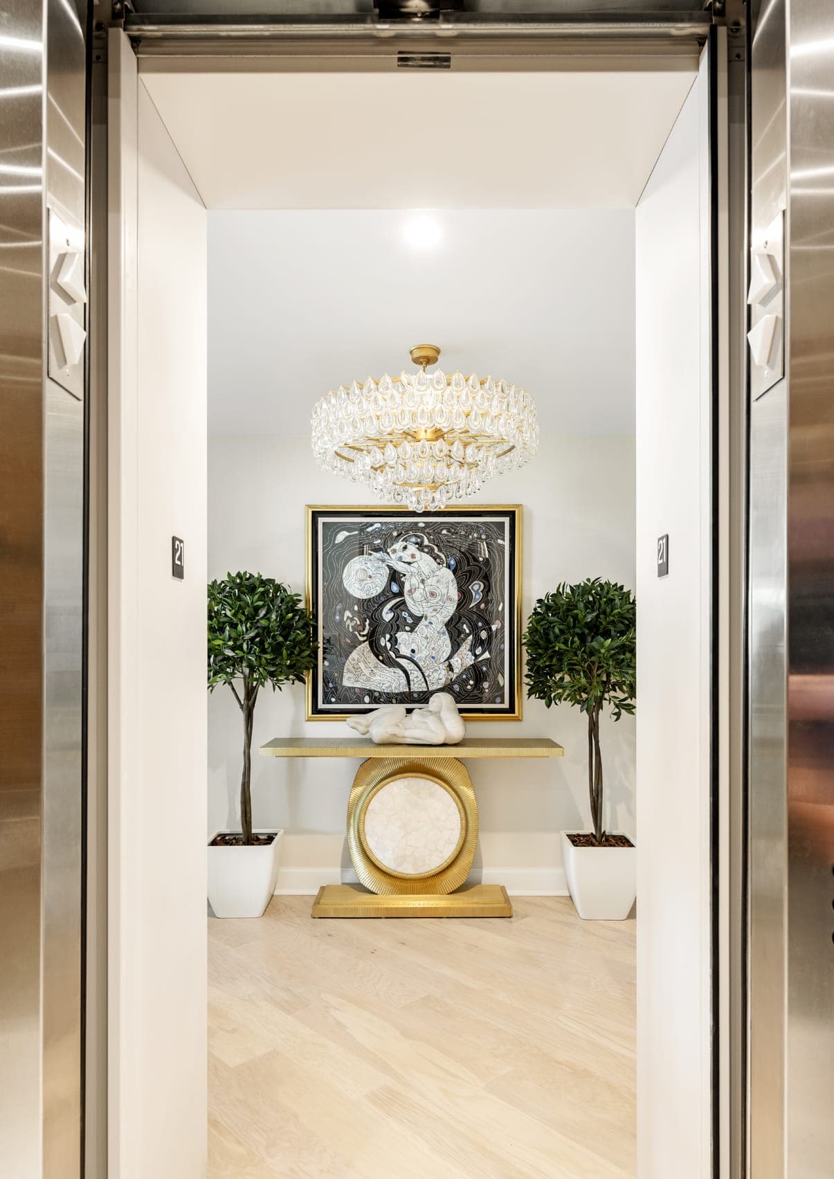 Lift Entrance To Condo Crystall Chandelier Gold Designer Table With White Stone Center Bay Bushes Abstract Dark Painting On Wall